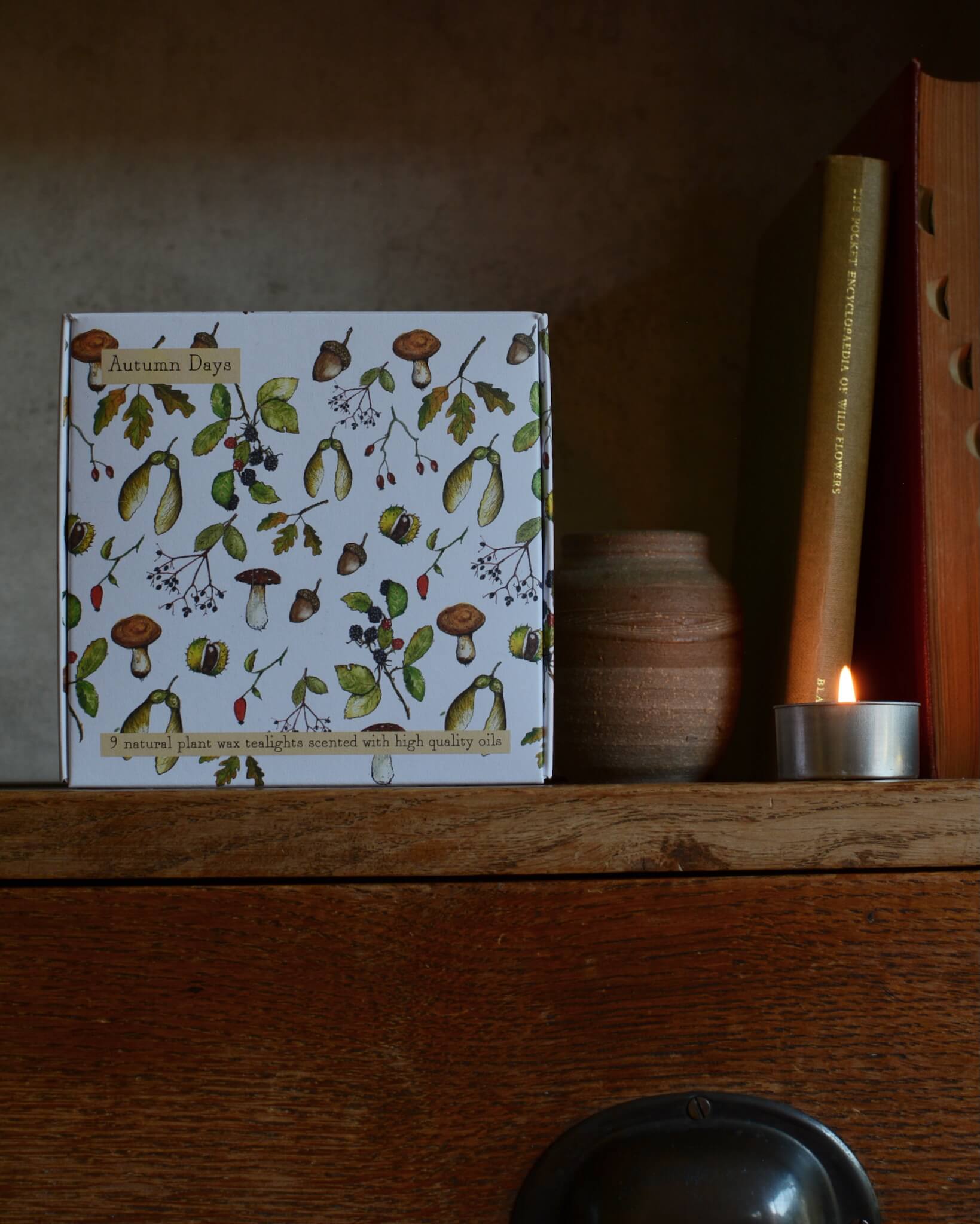 Autumn-scented tealights with nature-themed cover on a wooden shelf with a candle and decorative item.