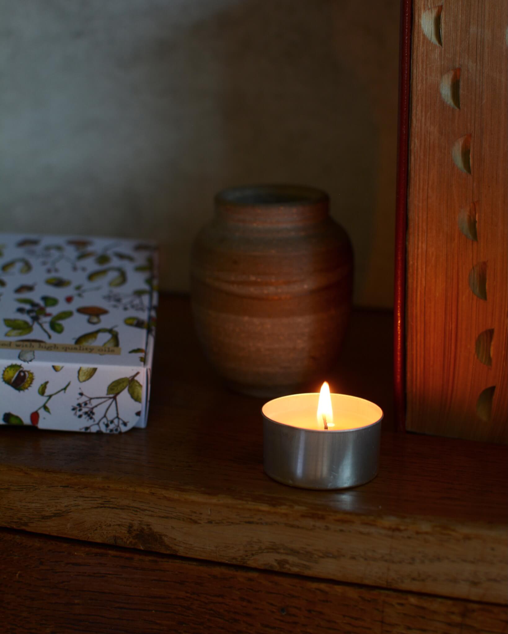 Tealight candle on a wooden surface with illustrated box and vase in the background