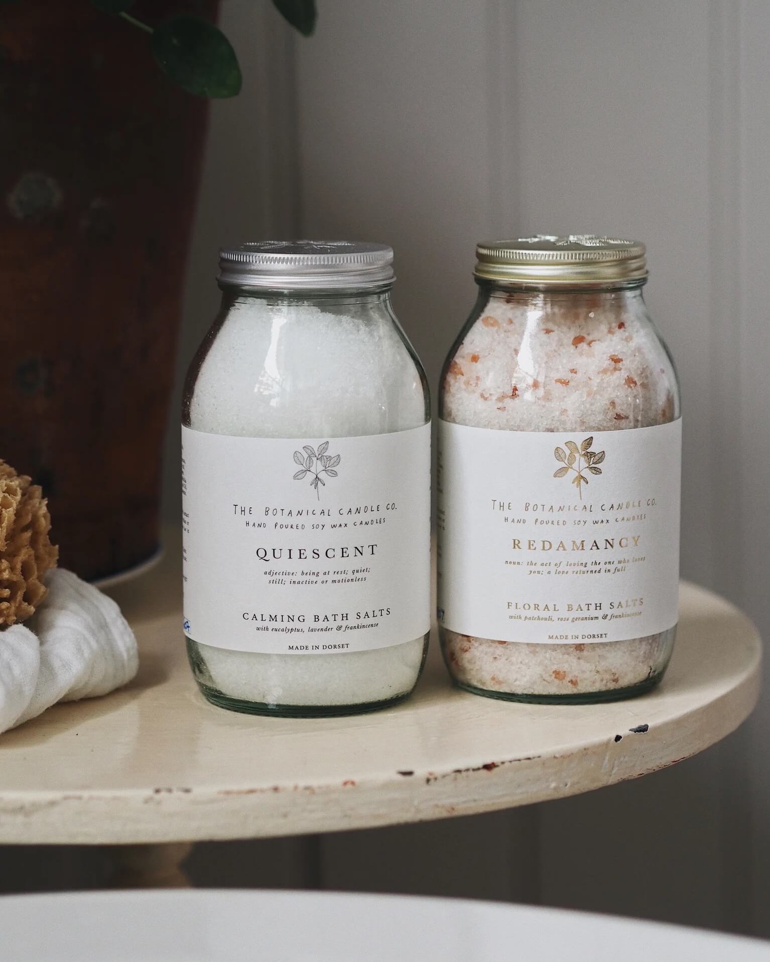 Two jars of bath salts labeled 'Quiescent' and 'Redamancy' on a wooden bathroom surface.