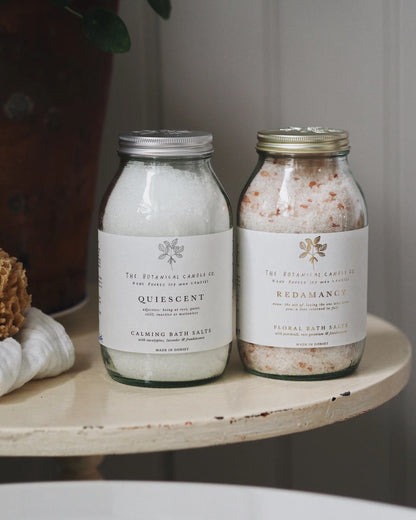 Two jars of bath salts labeled 'Quiescent' and 'Redamancy' on a wooden bathroom surface.