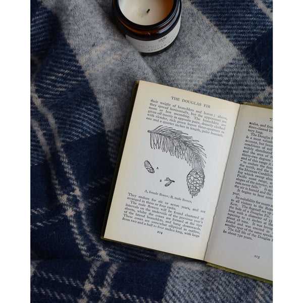 A woollen check blanket with a jar candle and open book depicting botanical illustration. 