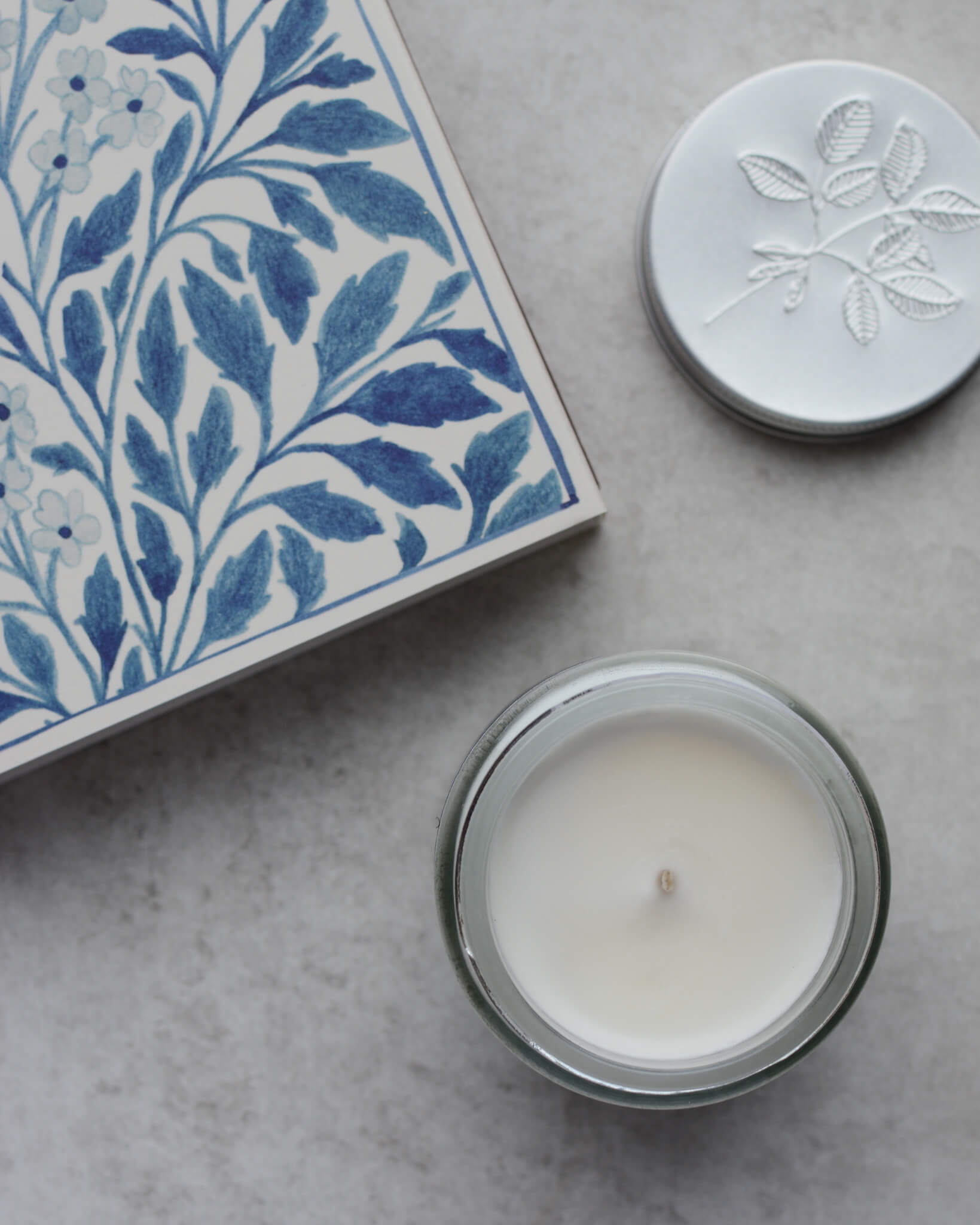 An open white candle with lid removed, next to a box of blue floral matches.