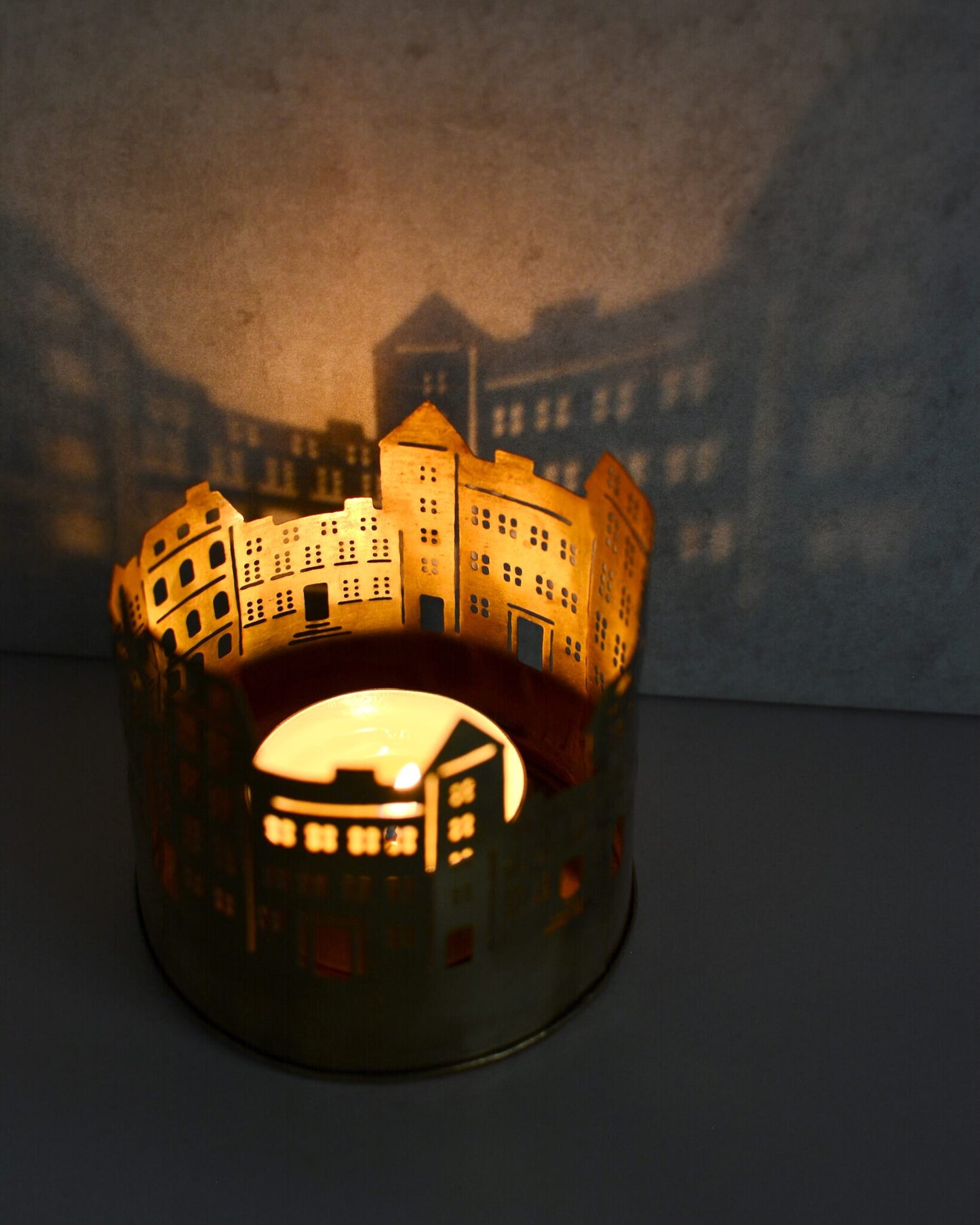 Brass tealight candle holder shaped like a cityscape, projecting townhouse shadows onto the wall behind.