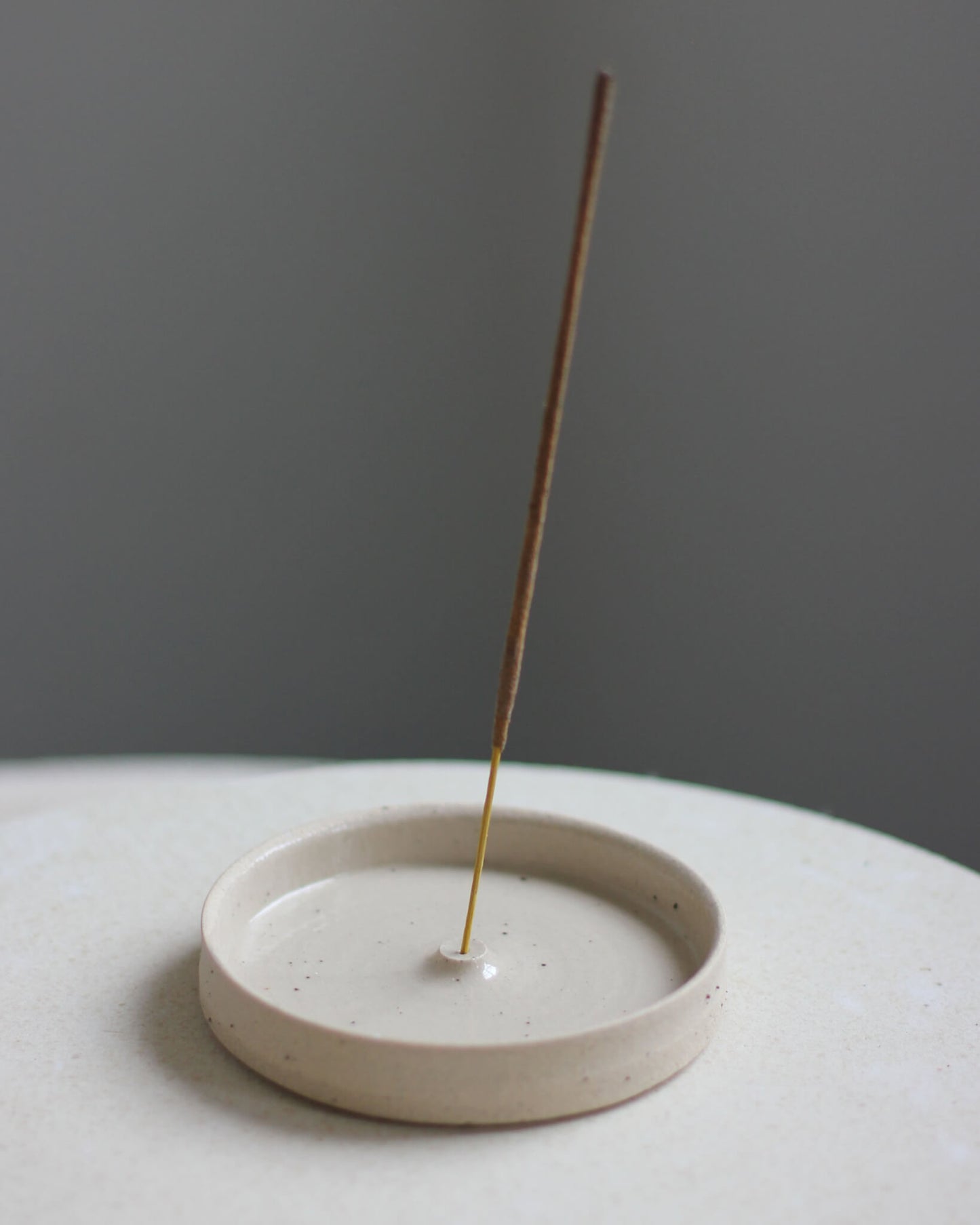 An oatmeal coloured ceramic hand-thrown incense holder, with an incense stick.