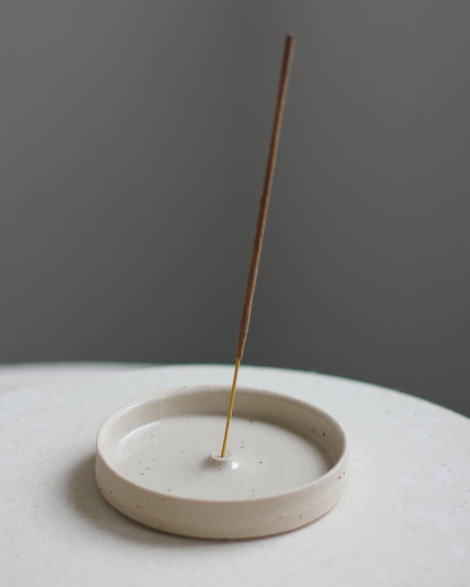 An oatmeal coloured ceramic hand-thrown incense holder, with an incense stick.