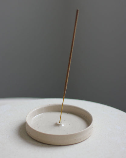 An oatmeal coloured ceramic hand-thrown incense holder, with an incense stick.