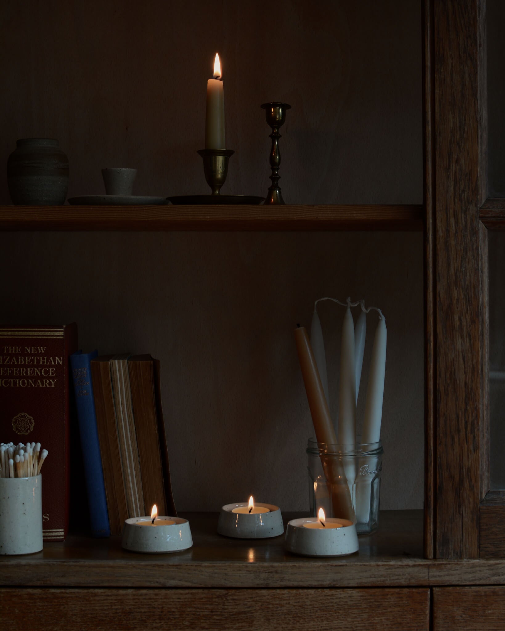 A collection of ceramic tea light holders, shown with lit beeswax tea lights on wooden shelves.