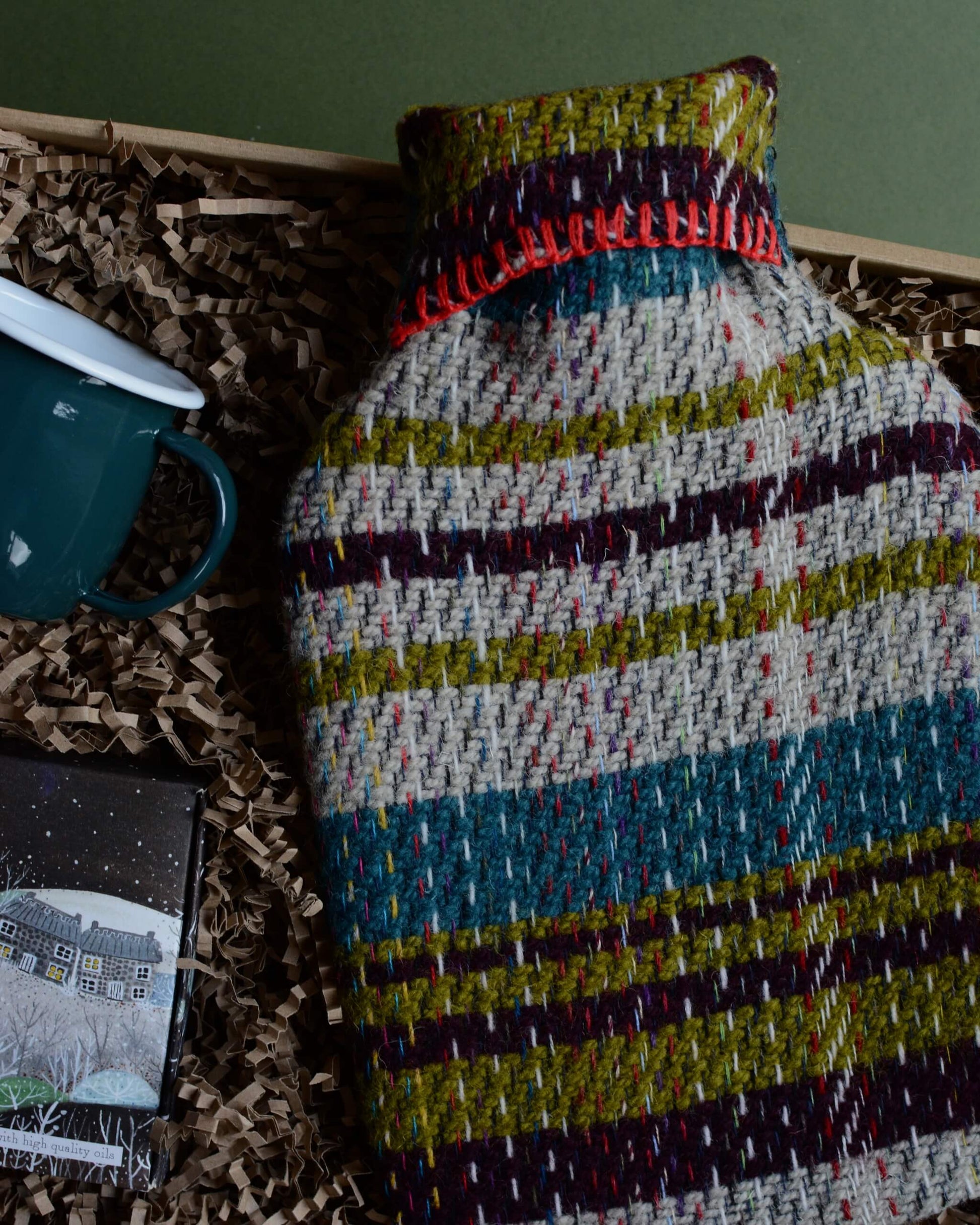 A wool hot water bottle, green enamel mug, and box of tealights in a gift box, shown on a green background.