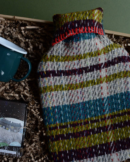 A wool hot water bottle, green enamel mug, and box of tealights in a gift box, shown on a green background.