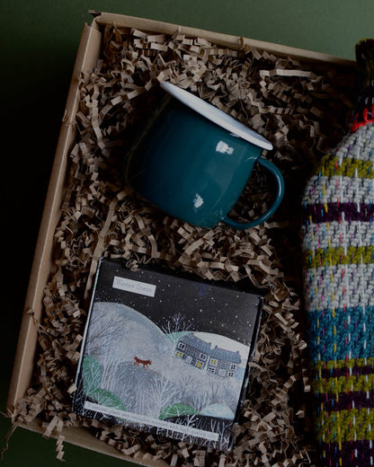 A wool hot water bottle, green enamel mug, and box of tealights in a gift box, shown on a green background.