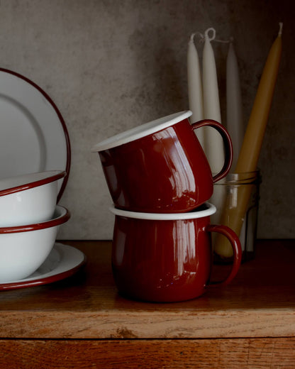 A pair of deep burgundy red enamel mugs, alongside other enamel tableware. 