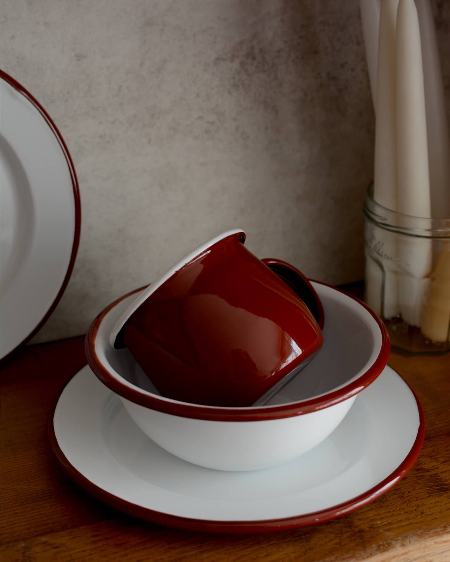 A deep burgundy red enamel mug, alongside other enamel tableware. 