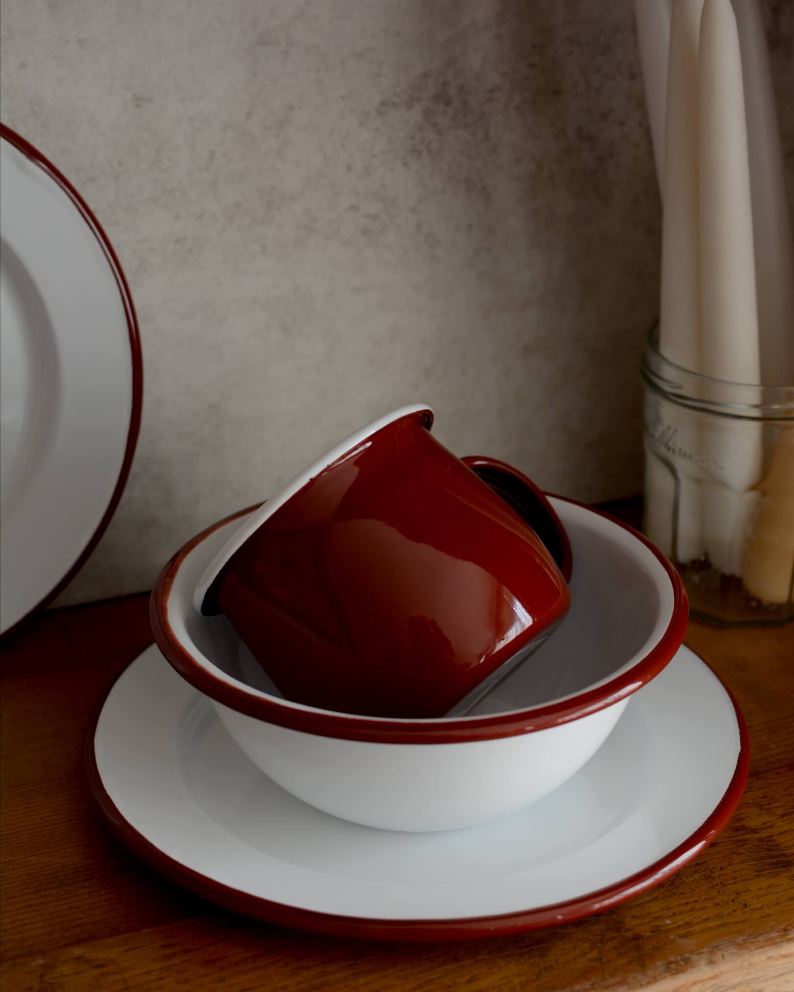 A deep burgundy red enamel mug, alongside other enamel tableware. 