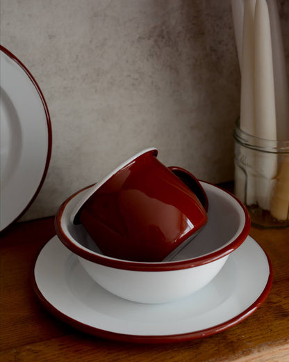 A deep burgundy red enamel mug, alongside other enamel tableware. 