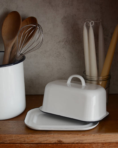 A white enamel butter dish with a navy blue rim, next to an enamel utensils holder and beeswax candles.