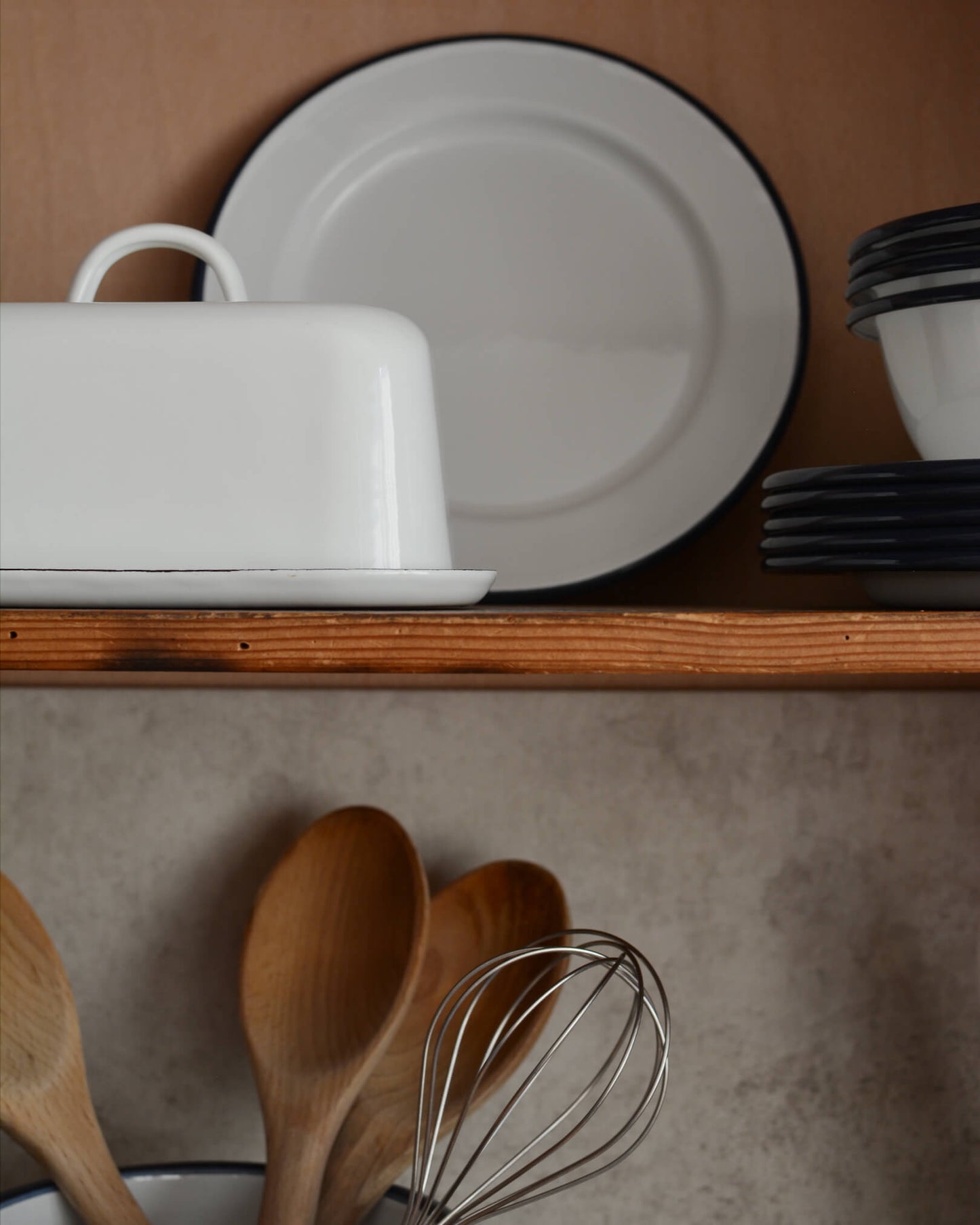 A white enamel butter dish with other enamel kitchenware on kitchen shelves.