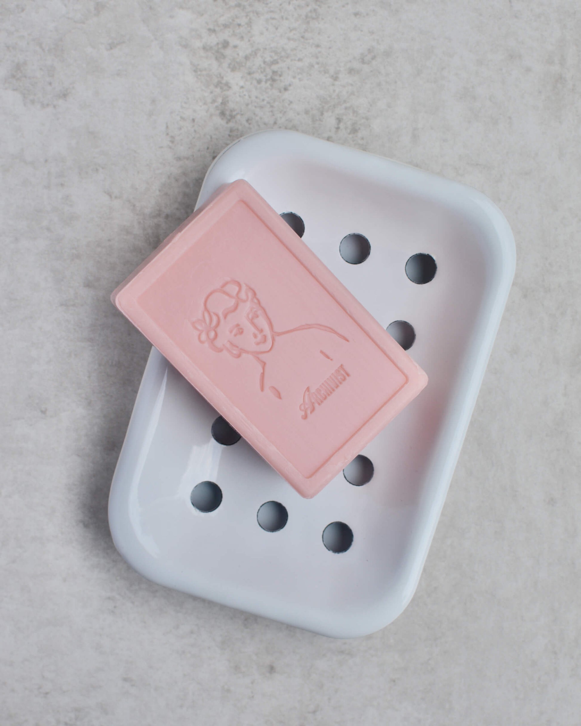 Pink soap bar with a brand logo on a white soap dish against a light grey background