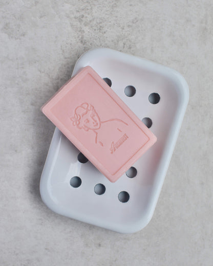 Pink soap bar with a brand logo on a white soap dish against a light grey background