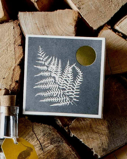 Fern print design matchbox, placed on stacked wooden logs.