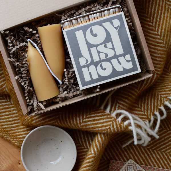 A gift box containing beeswax candles and a box of matches. Next to a ochre yellow wool blanket.