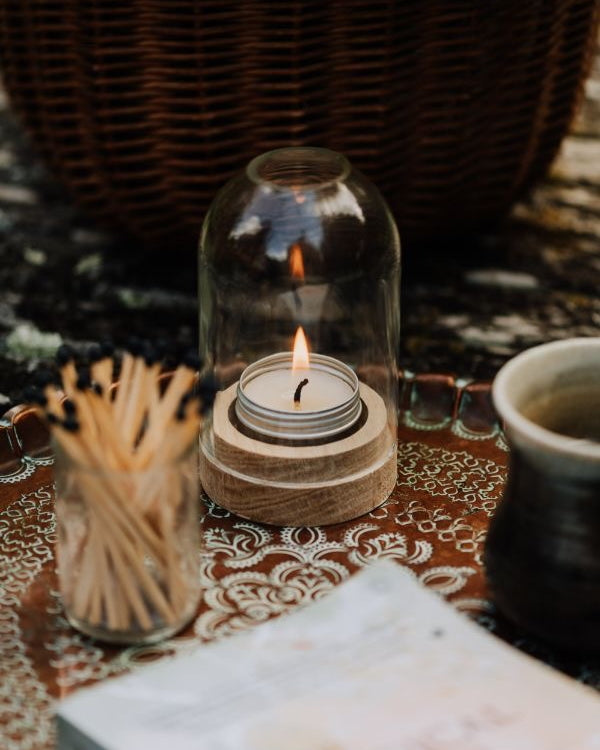 Candle in a glass dome lantern on a patterned tablecloth with matches and a cup.