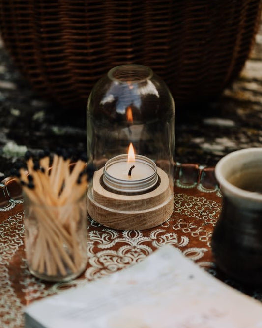 Candle in a glass dome lantern on a patterned tablecloth with matches and a cup.