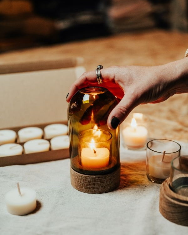 Hand holding a glass lantern with a lit candle, surrounded by other candles on a soft surface.