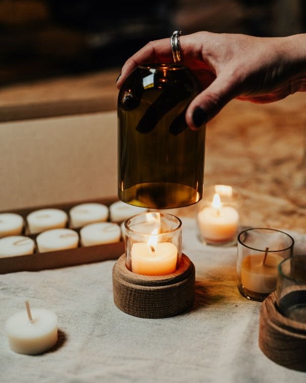 Hand holding a green glass candle lantern over a small candle, surrounded by other candles on a wooden surface.