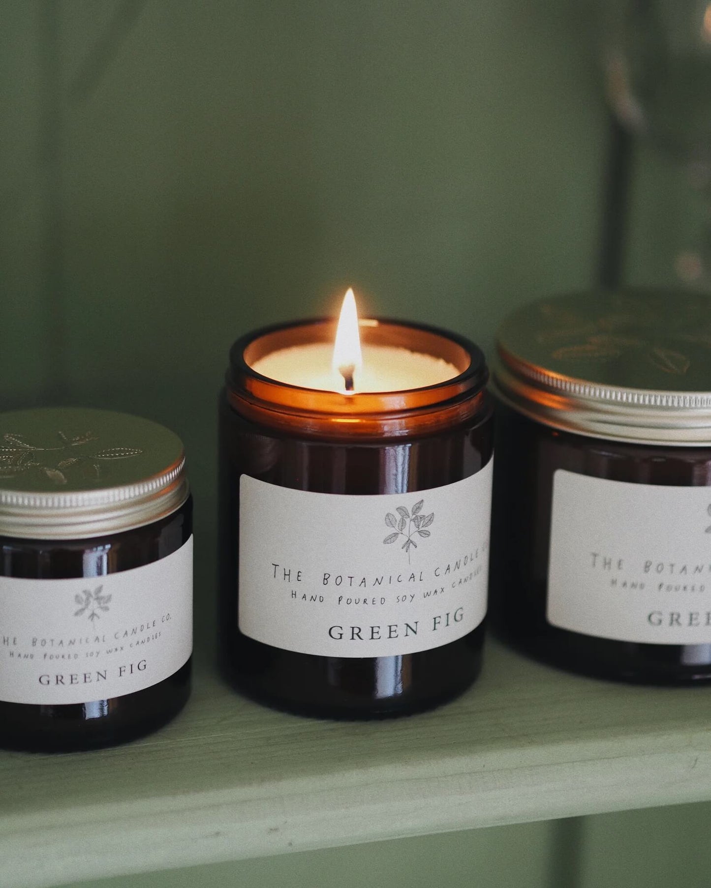 Candle labeled 'Green Fig' with two jars on a shelf against a green background