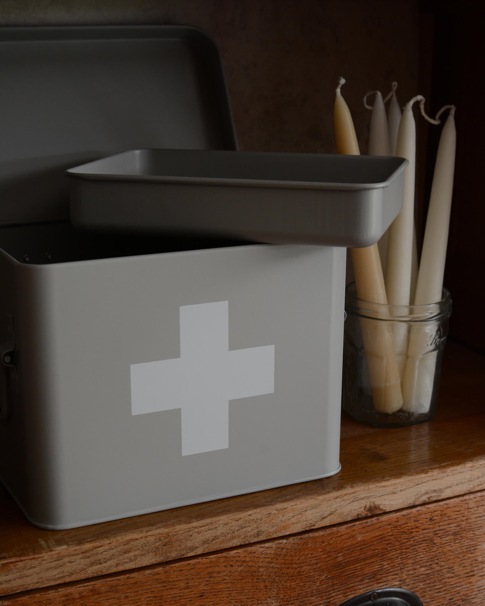 A matte grey metal first aid box with a white cross symbol on the front, shown open with the inner tray on top, placed on a wooden surface.