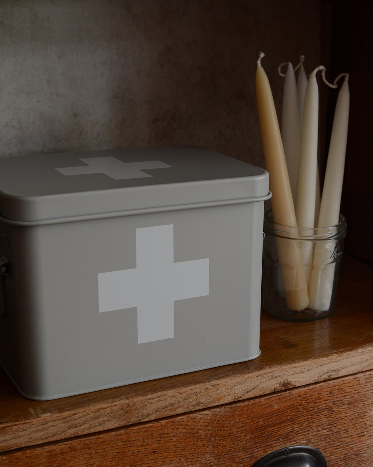 A grey metal first aid tin with a white medical-style cross on the lid and front, placed on a wooden surface next to a glass jar holding taper candles.