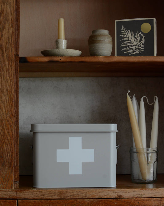 A light grey metal first aid box with a white cross on the front, displayed on a wooden shelf next to candles and decorative objects.