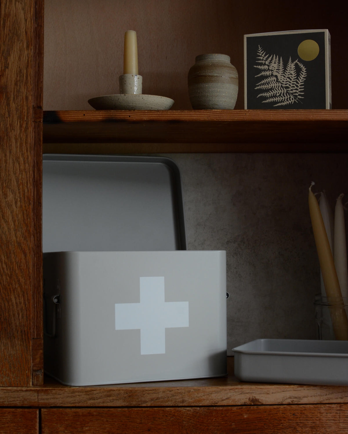 A light gray metal first aid box with a white cross symbol on the front, displayed on a rustic wooden shelf next to a ceramic vase, candles, and a framed print.