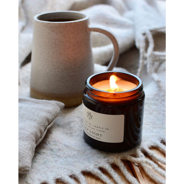 Half Light Scented Soy Candle in amber jar on a neutral beige blanket, next to a ceramic coffee mug.