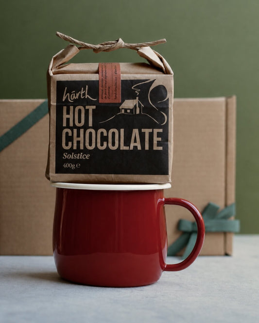 A packet of Harth hot chocolate on top of a red enamel mug, in front of a kraft gift box with a green ribbon.