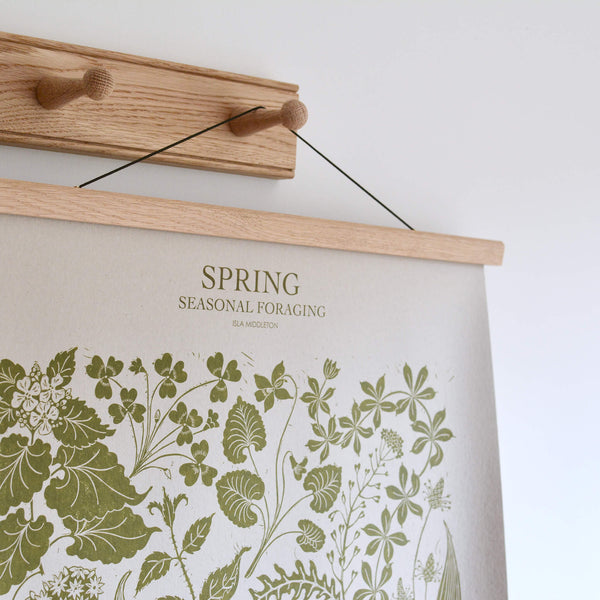 Botanical 'Spring' print with green ink on pale grey paper, hanging from an oak poster frame on a set of oak wall hooks.