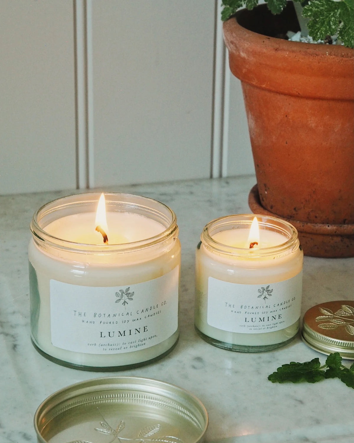 Two lit Lumine scented candles by The Botanical Candle Co. in clear glass jars with white labels, displayed on a marble surface next to a terracotta planter.