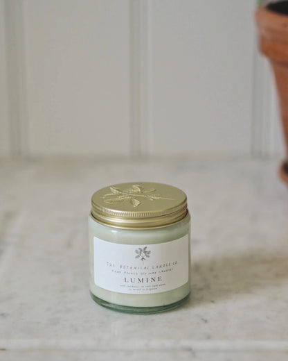 A glass jar candle with a metallic gold lid sits on a light marble surface against a soft, blurred white panelled background. The label reads 'The Botanical Candle Co.