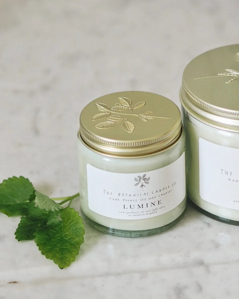 Two Lumine candles with gold lids sit on a light marble surface. The label reads "The Botanical Candle Co.