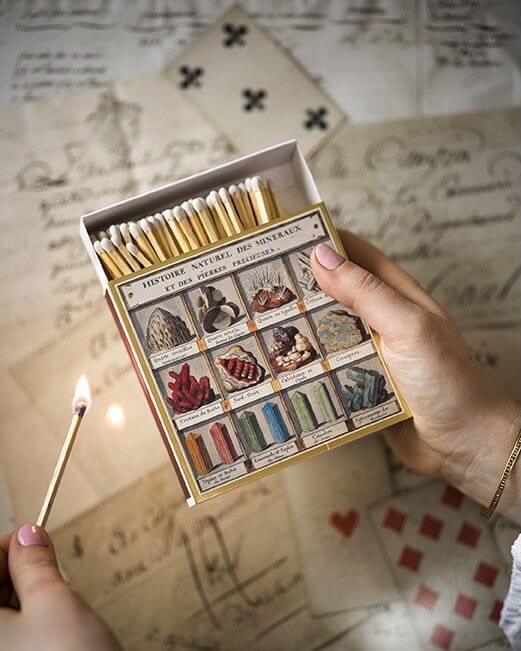 Matchbox with mineral illustrations held open by a hand with a lit match, on a textured surface.
