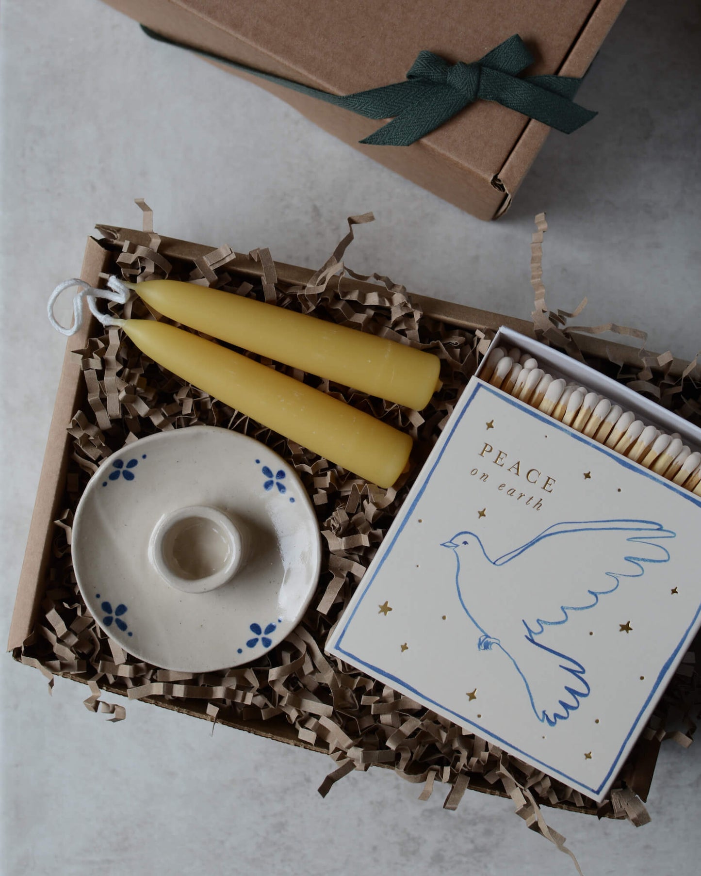 A white matchbox, with a dove illustration and 'peace on earth' lettering in a gift box, with a pair of beeswax candles and a white and blue pottery candle holder.