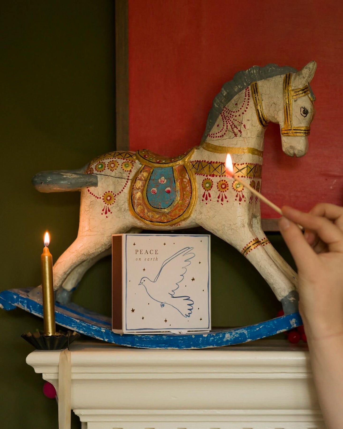Decorative rocking horse with candles and a illustrated matchbox on a mantelpiece, against a red wall.