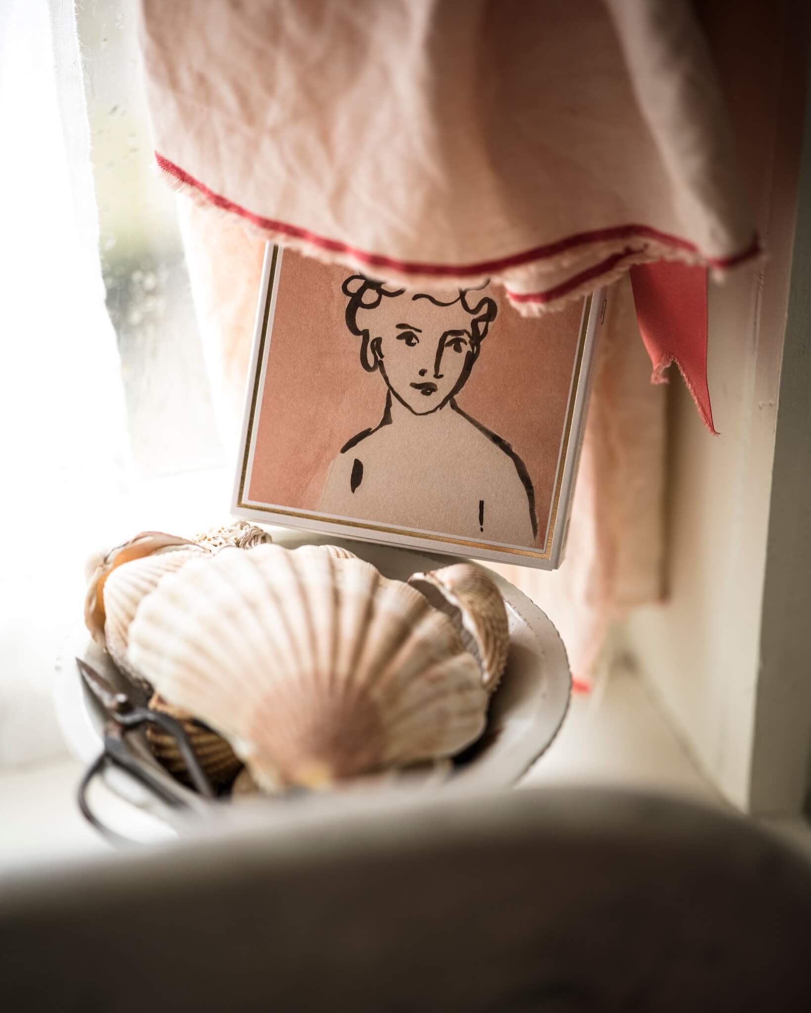 A pink matchbox featuring an illustrated portrait of a woman, shown on a bathroom shelf.