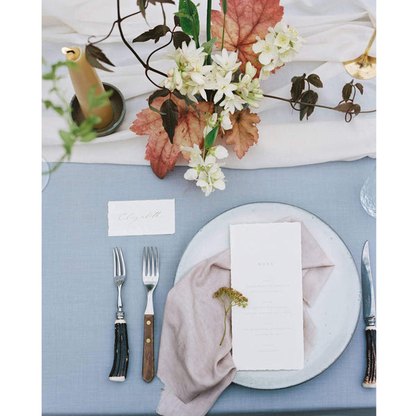 A large beeswax candle on top of a table with an informal flower arrangement and cutlery.