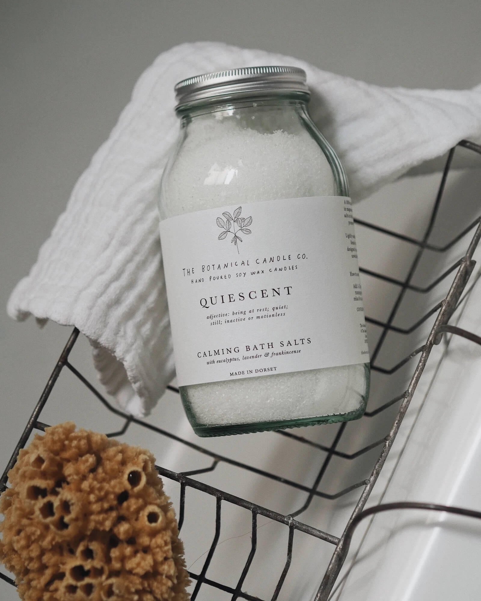Jar of bath salts labeled 'Quiescent' on a wire rack with a towel and sponge.