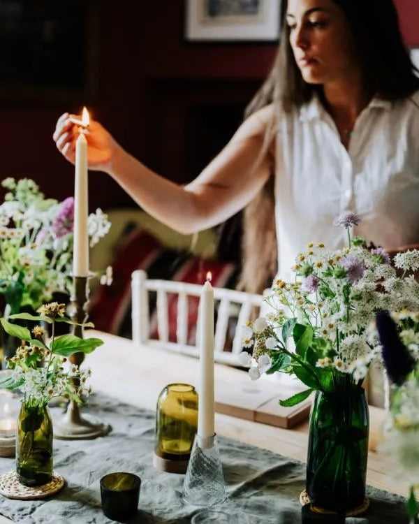A woman setting a dinner table and lighting long taper candles in glass candle holders.