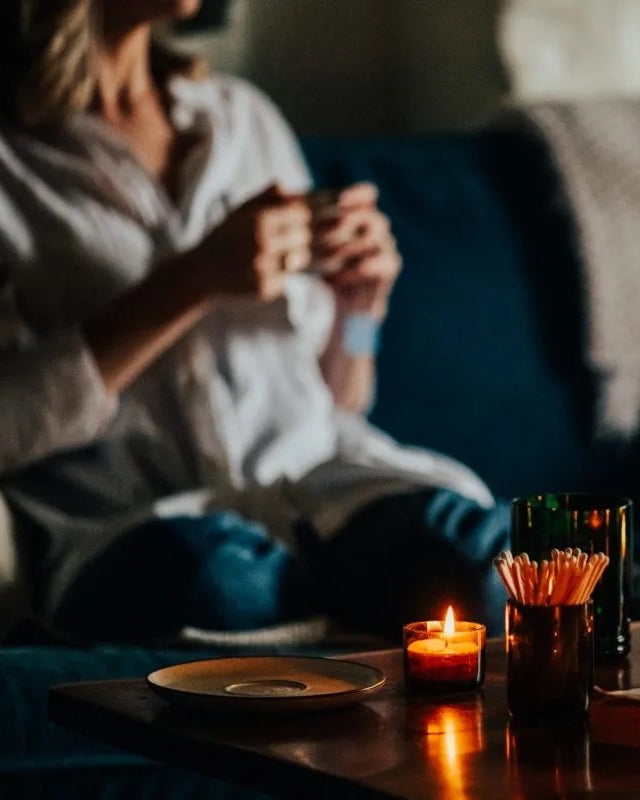A candlelit scene with candles and matches on a coffee table in front of a blurred background, showing a figure sat on a sofa.
