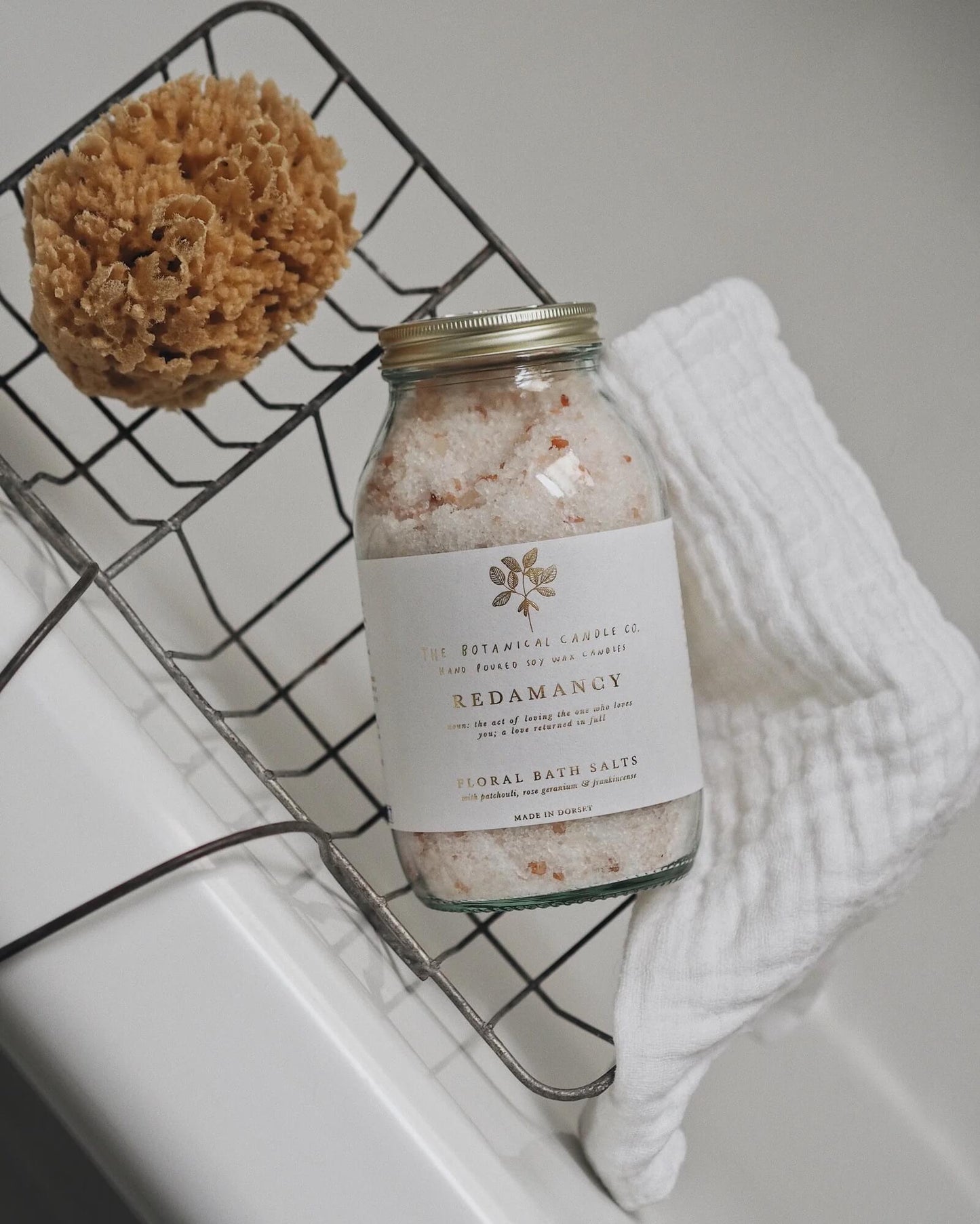 Bottle of bath salts labeled 'Redamancy' on a wire rack with a sponge and towel.