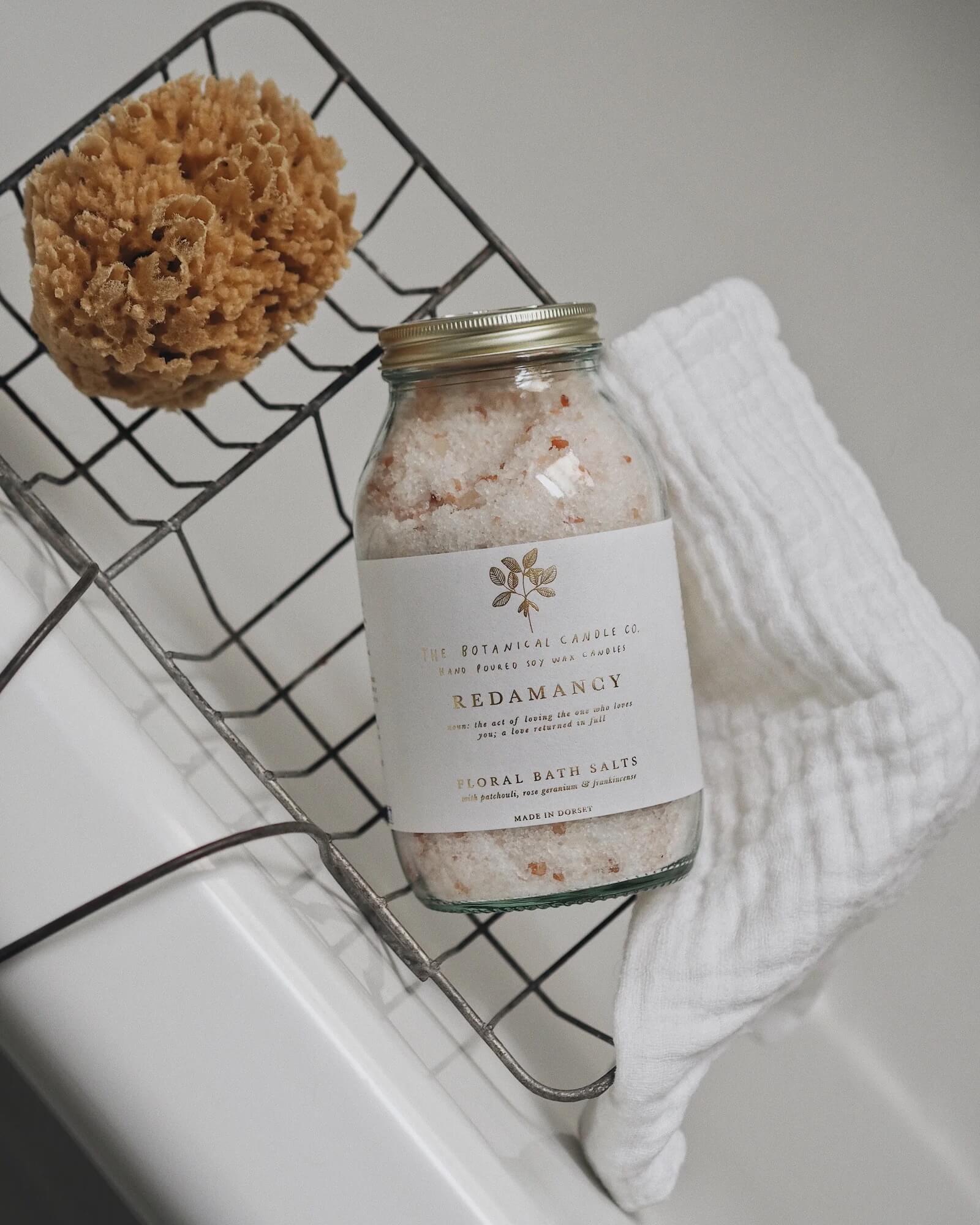 Bottle of bath salts labeled 'Redamancy' on a wire rack with a sponge and towel.