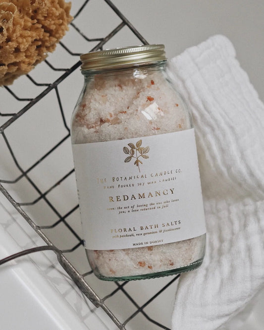 Bottle of floral bath salts labeled 'Redamancy' on a wire rack with a white towel.